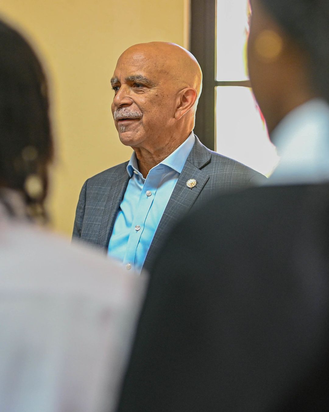 A bald man with a grey moustache, wearing a grey suit jacket and a light blue shirt, stands and speaks to a group of attendees whose backs are to the camera.