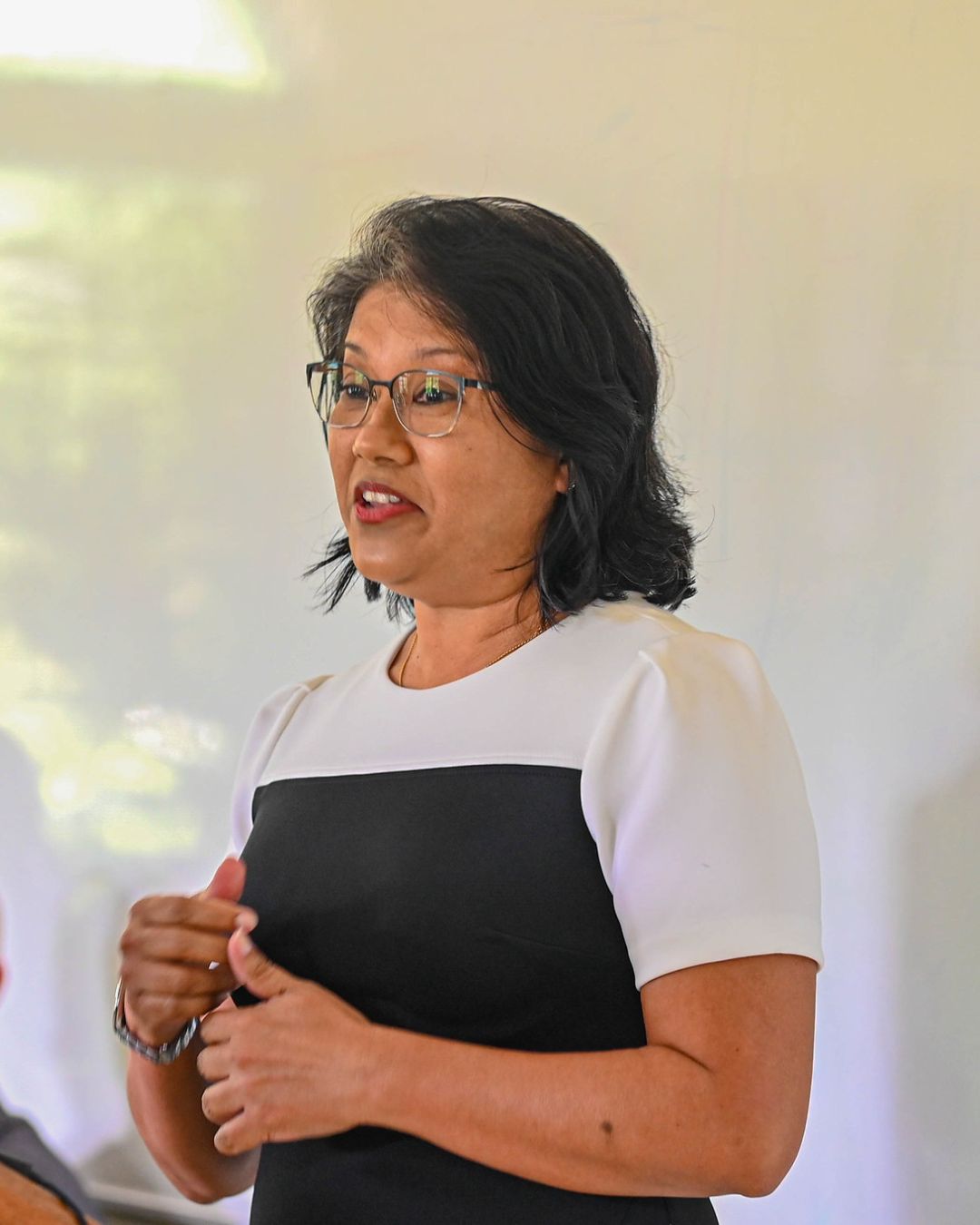 A woman wearing glasses and a black-and-white colour-blocked dress stands at a wooden podium with a laptop, speaking to an audience.