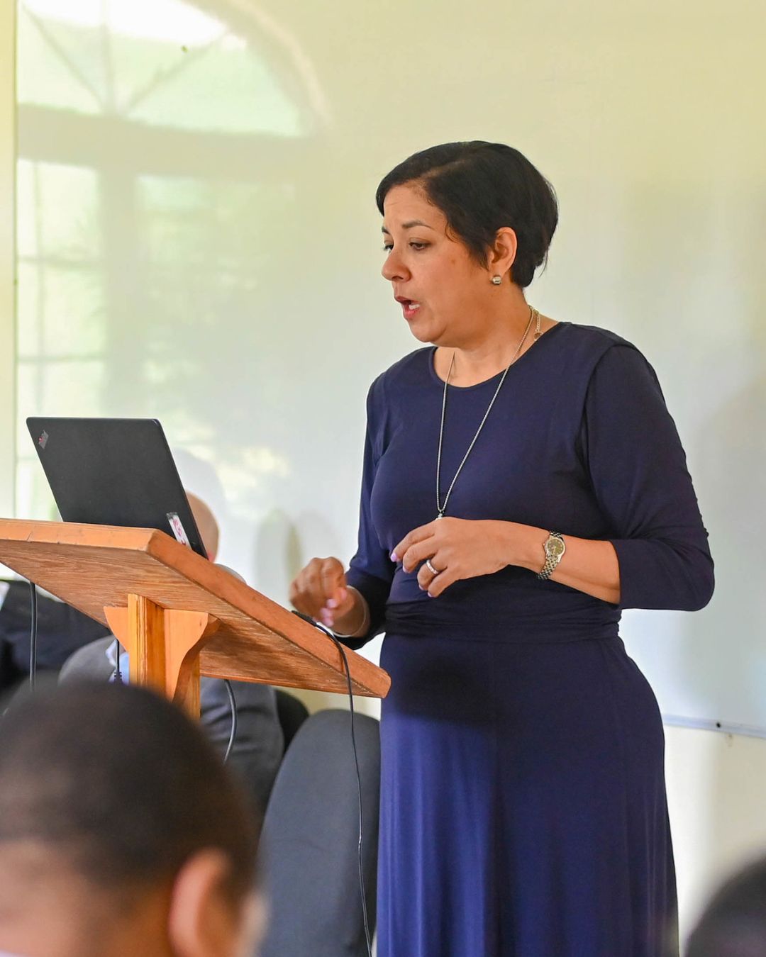 A woman with short dark hair, wearing a long dark blue dress and a silver necklace, stands behind a wooden podium with a laptop, gesturing with her right hand as she speaks.