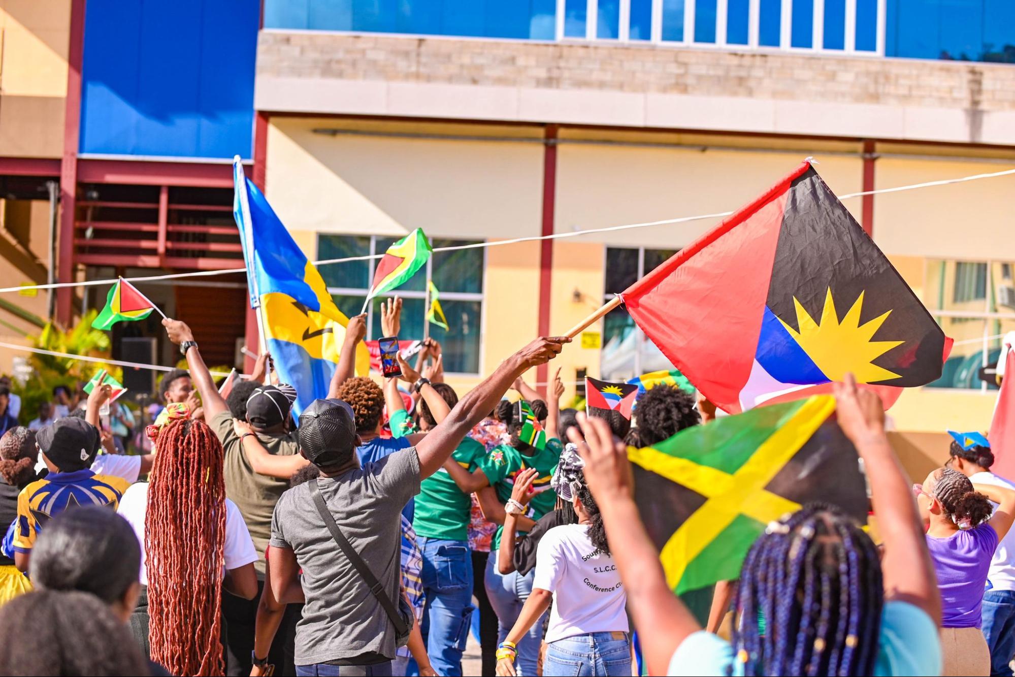 A large crowd of students seen from behind, enthusiastically waving various Caribbean flags, including those of Antigua, Jamaica, and the Bahamas at ISW 2026