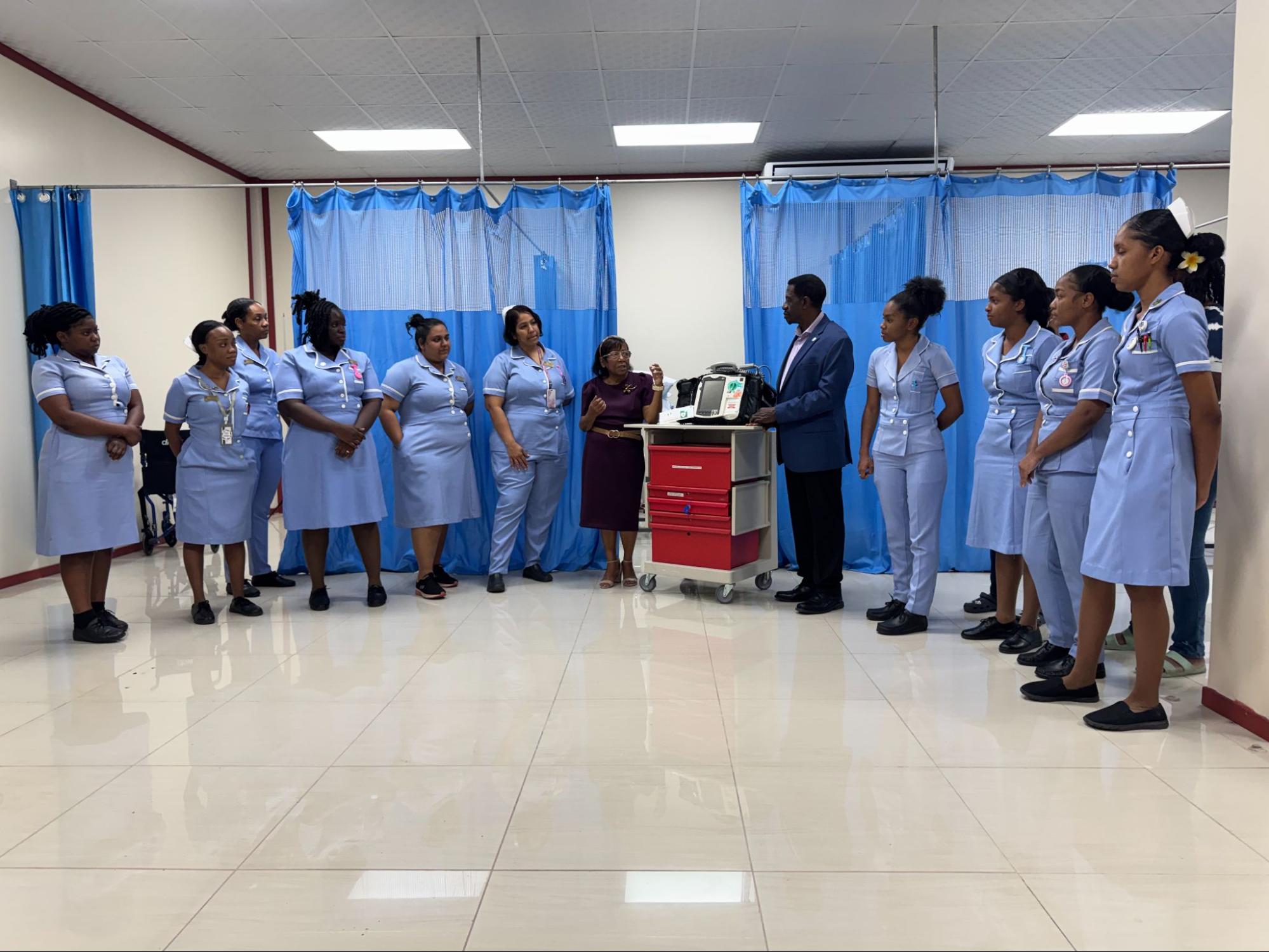 A wide view of nursing students gathered around Dr. Wilson and Dr. Woolford near a red medical cart with a defibrillator inside the lab, partitioned by blue curtains