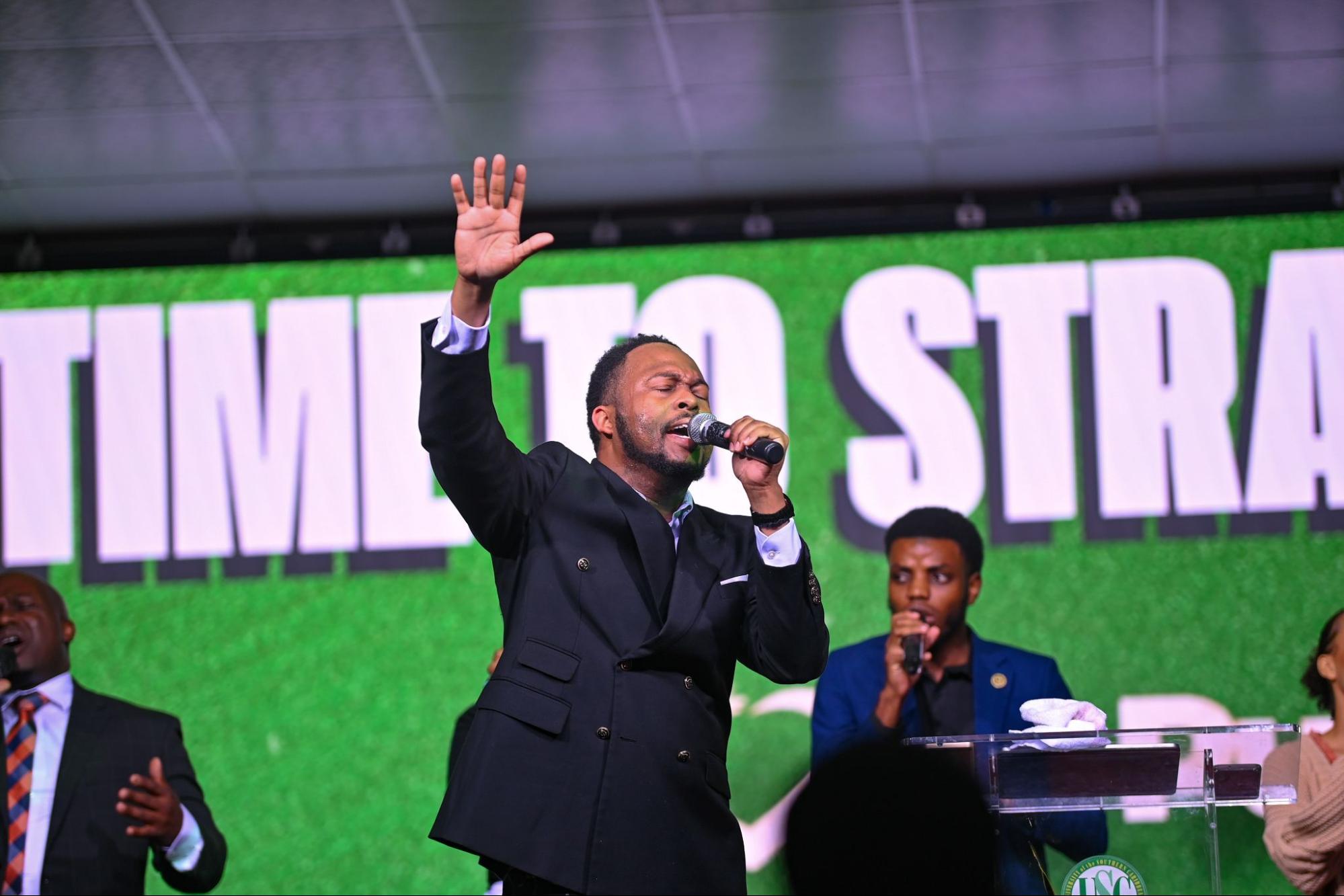 Pr. Jovan Whyte in a black double-breasted suit speaking passionately into a microphone with his right hand raised in worship.