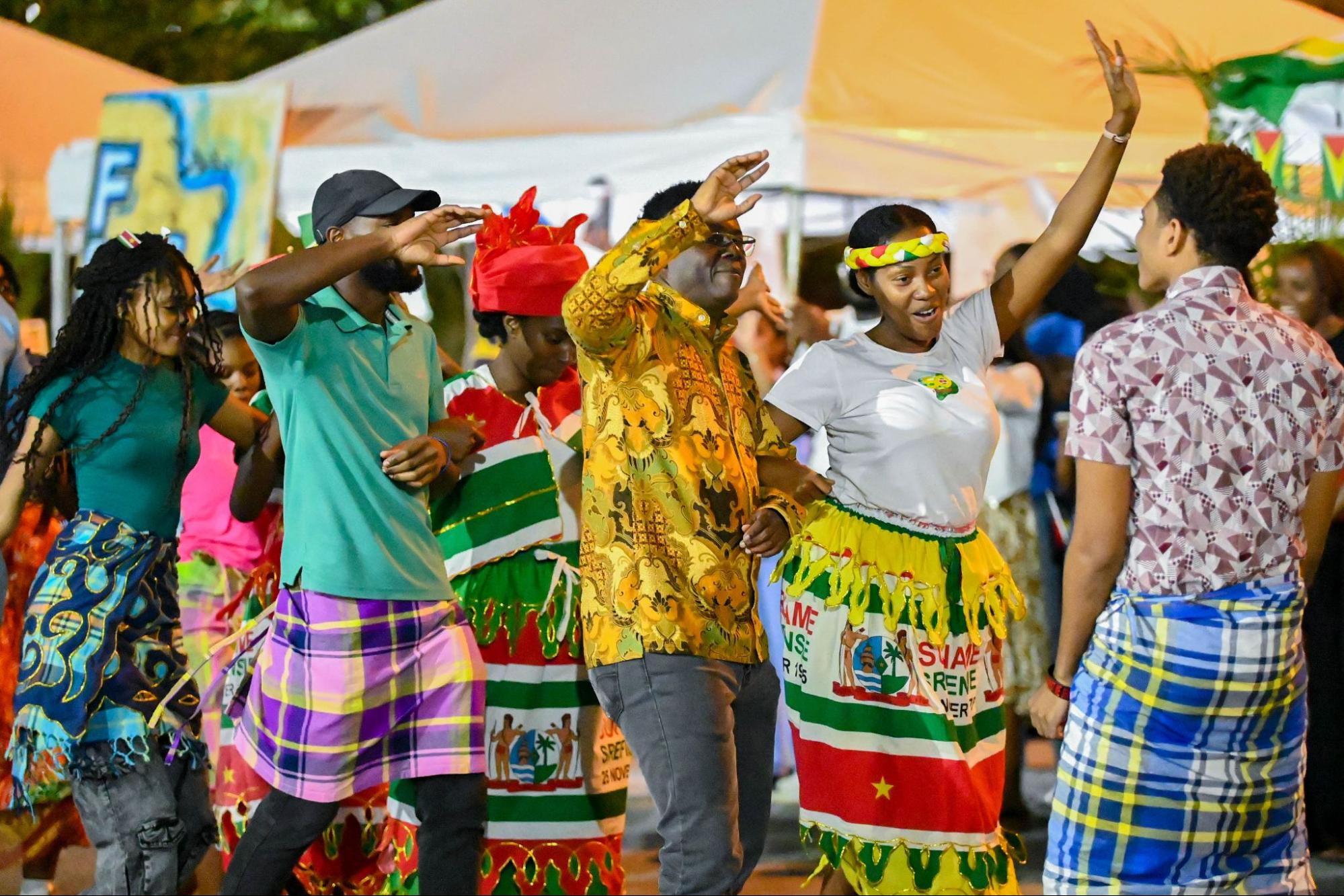 A group of students wearing traditional, brightly coloured Surinamese clothing dance together joyfully at an outdoor evening event