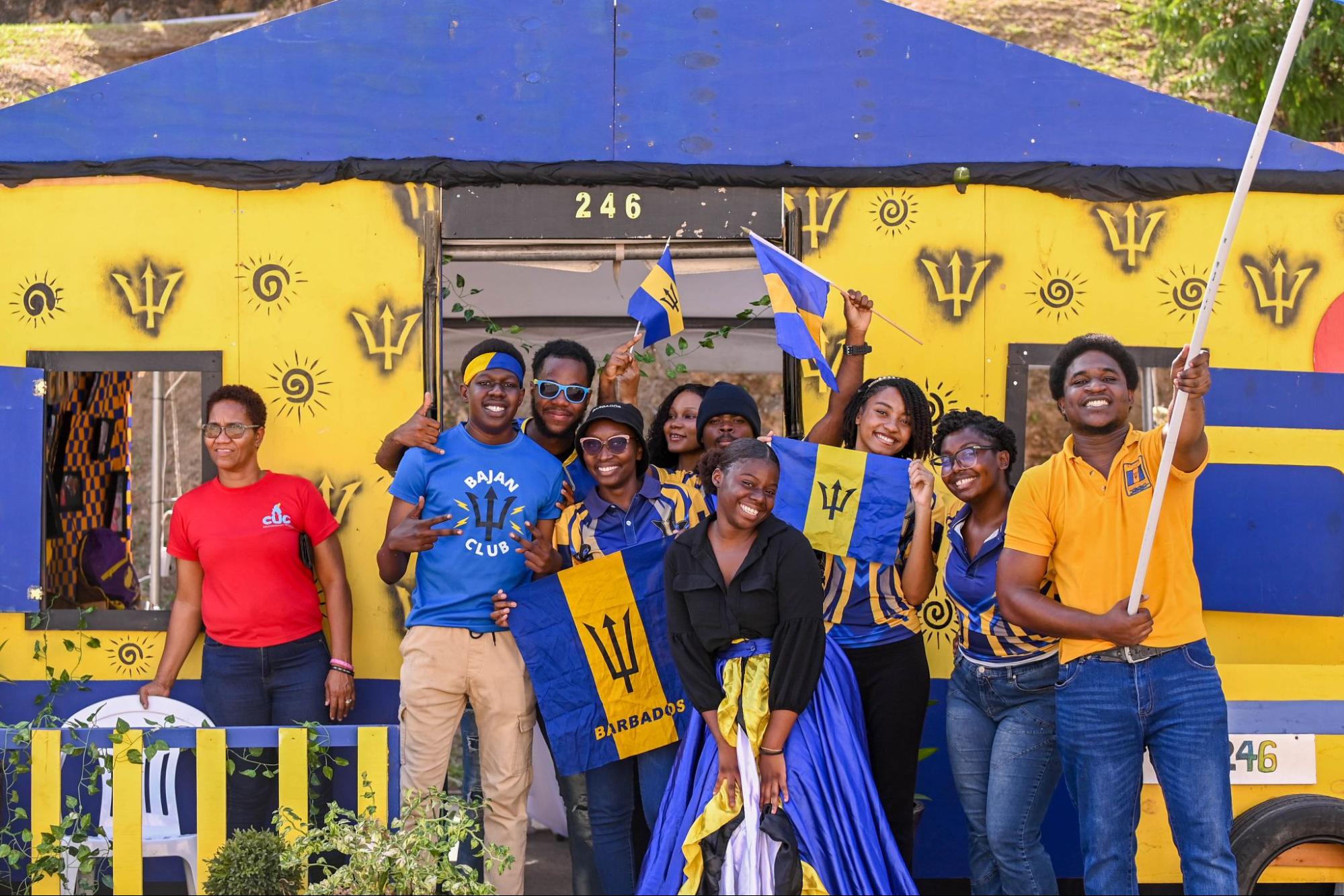 A group of smiling students pose with Barbadian flags in front of a blue and yellow wooden booth decorated with tridents and the number 246