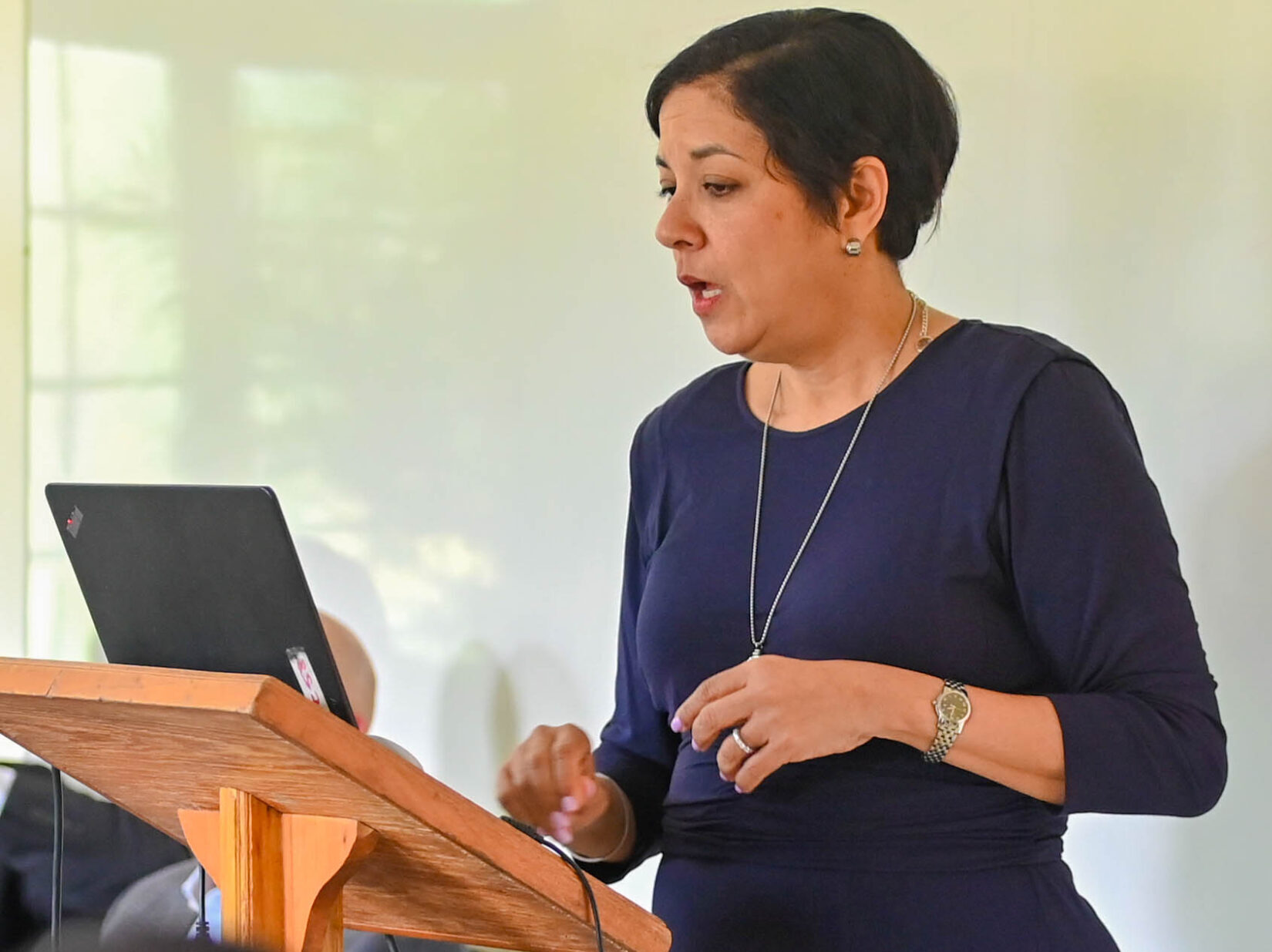 A woman with short dark hair, wearing a long dark blue dress and a silver necklace, stands behind a wooden podium with a laptop, gesturing with her right hand as she speaks.