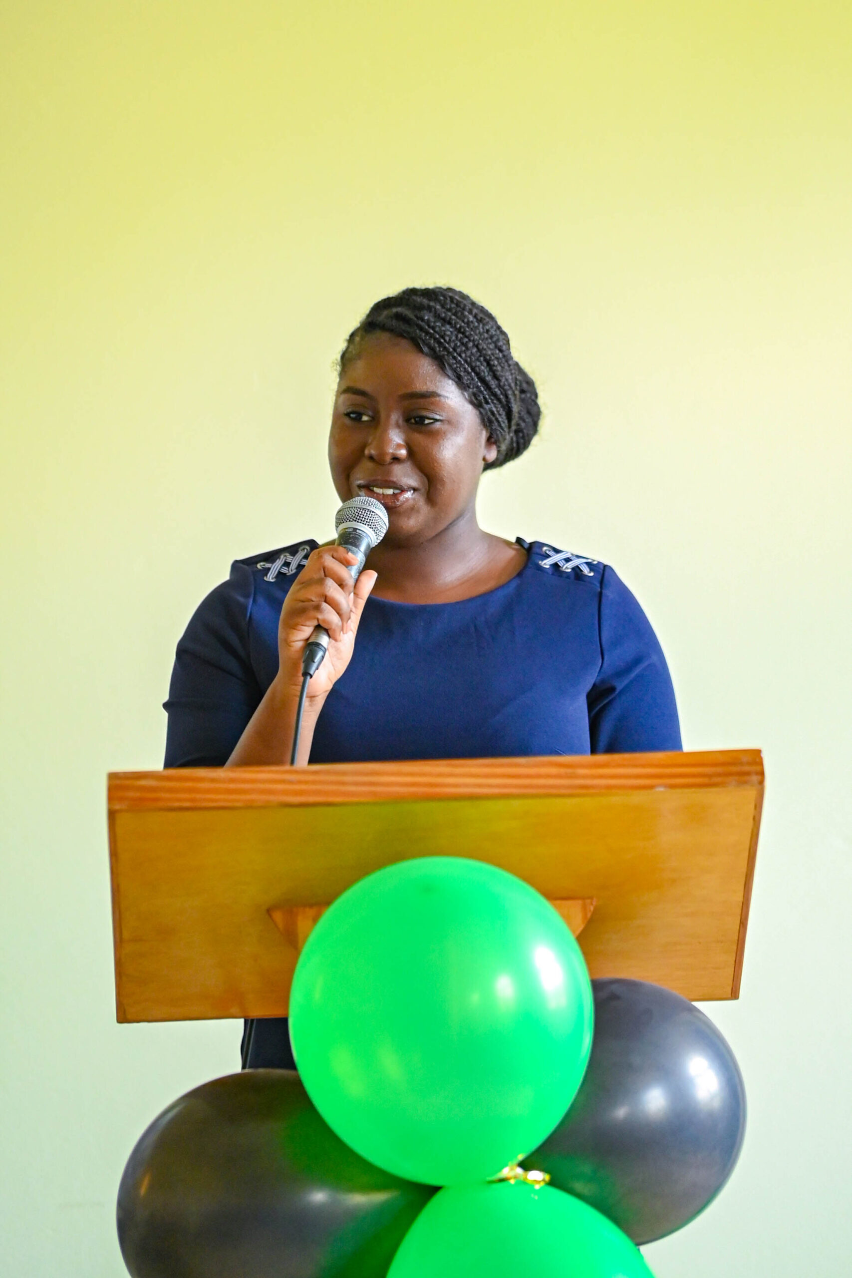 Vice Chair Ayandi Delpeche speaks into a microphone at a wooden podium, with a projection screen visible in the background