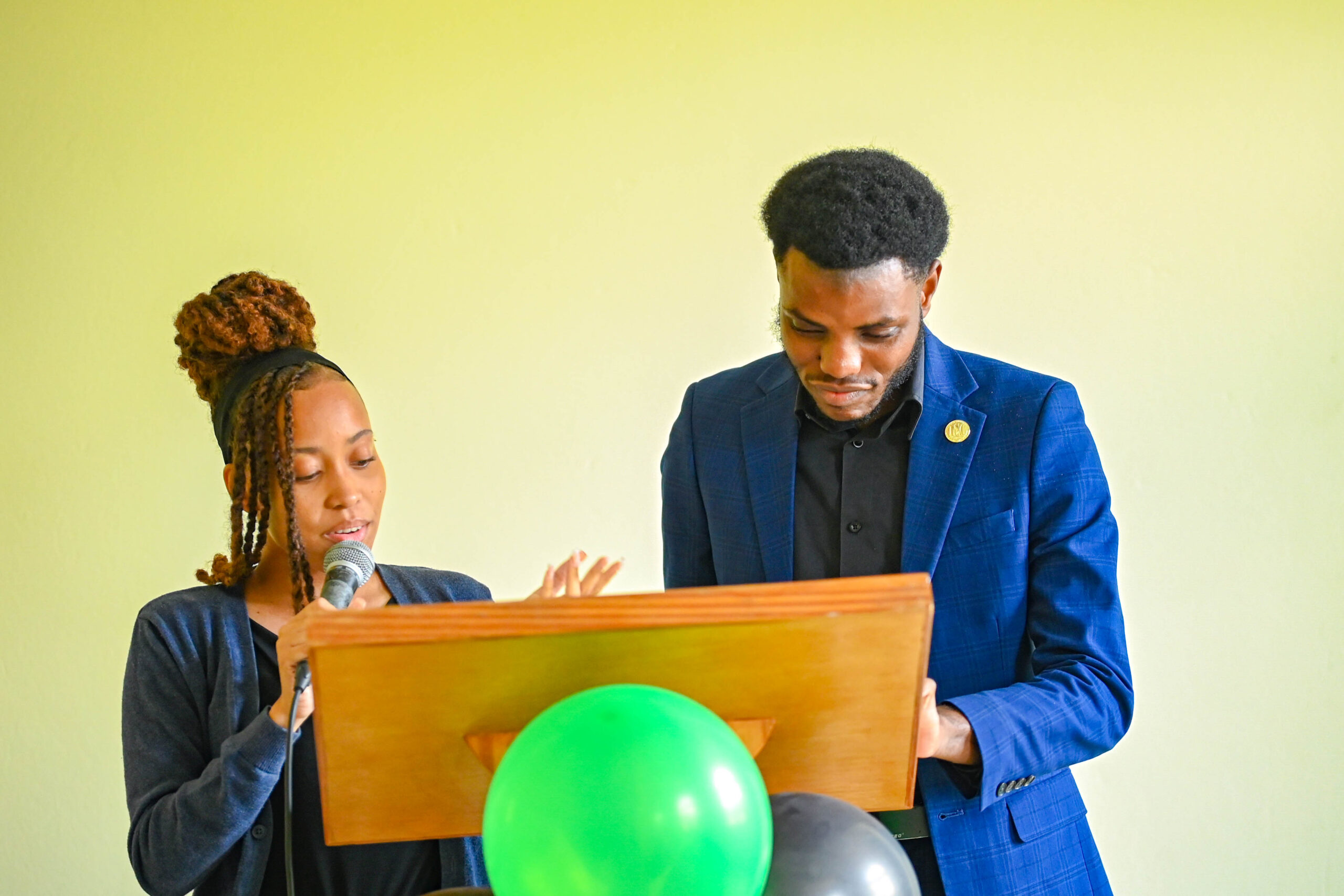Dayna-Marie Nichols speaks into a microphone at a wooden podium while Juhmaul Belfon stands beside her reviewing notes.