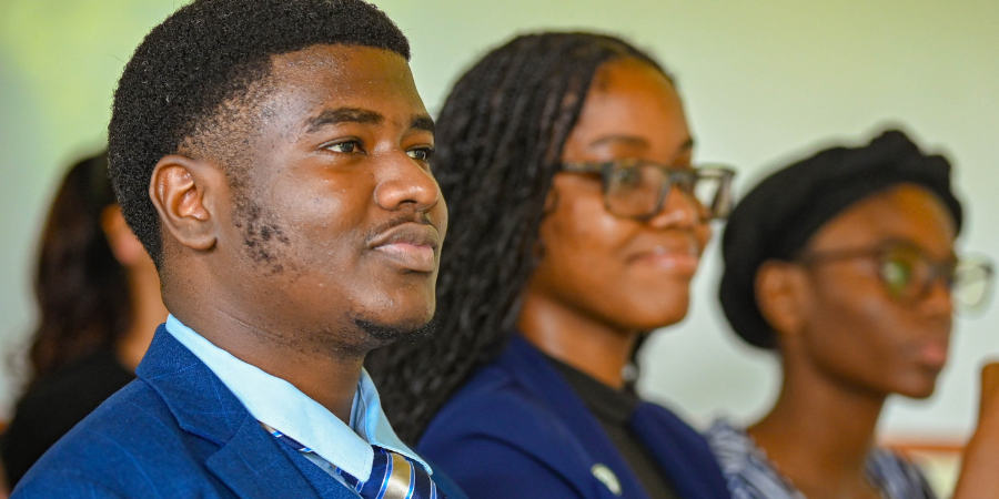 A young man in a blue suit and striped tie sits attentively next to a young woman wearing glasses at a formal meeting.