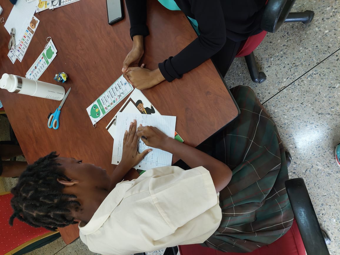 An overhead view of a San Juan North Secondary School student writing on lined paper while referencing a vocabulary card during a one-on-one literacy intervention session with a University of the Southern Caribbean mentor.
