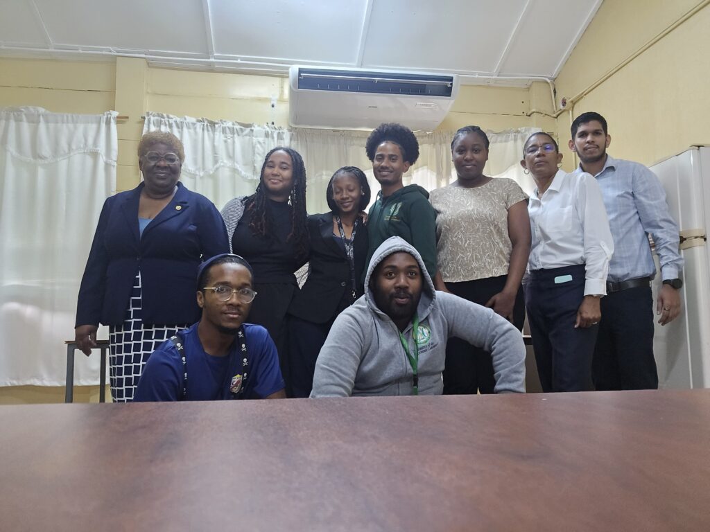 A group portrait of diverse University of the Southern Caribbean pre-service teachers, student leaders, and educational faculty members standing and sitting together in a classroom at San Juan North Secondary School.