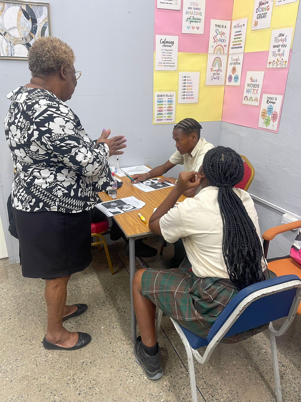 An educator from the University of the Southern Caribbean provides guidance to two seated San Juan North Secondary School students in a classroom decorated with growth mindset and calming strategy educational posters.