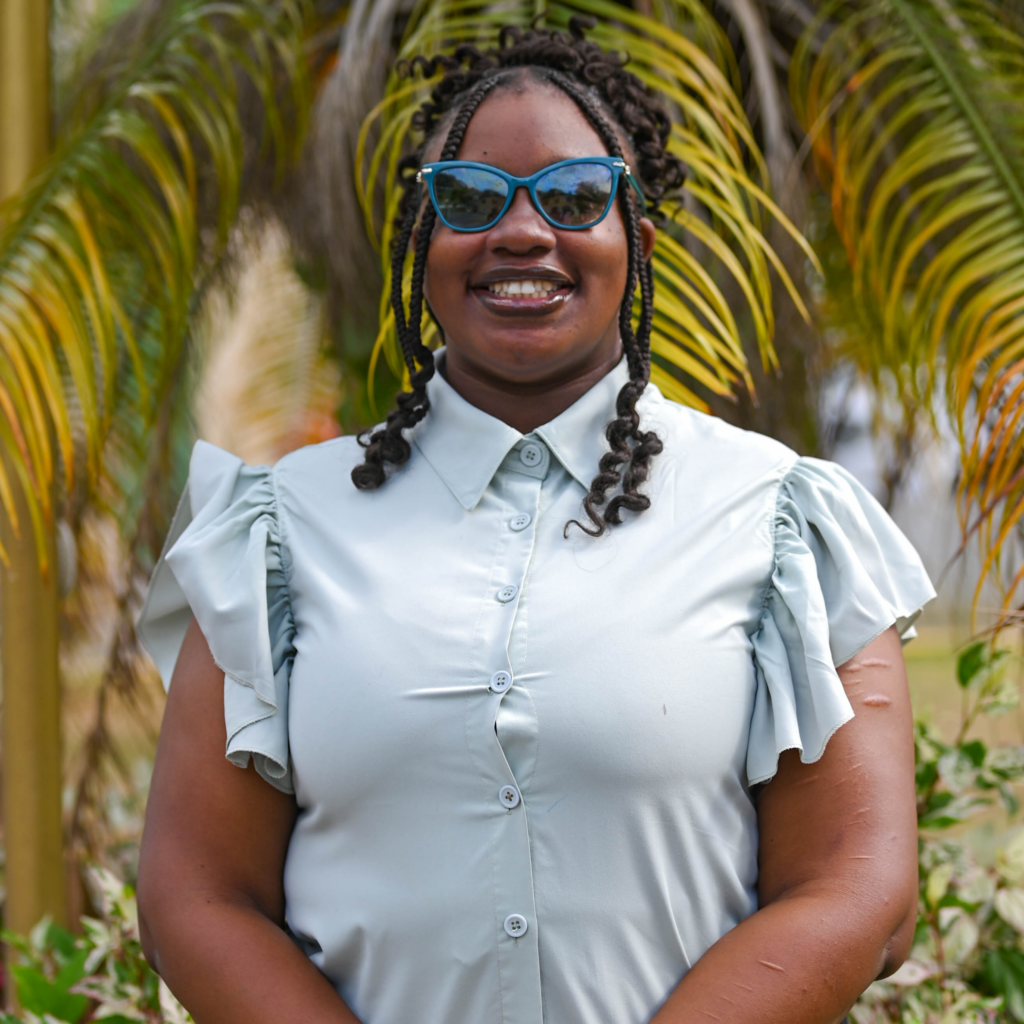 A portrait of a smiling woman with braided hair and blue-framed sunglasses, wearing a light-blue button-down blouse with ruffled sleeves, standing in front of lush palm leaves and greenery.