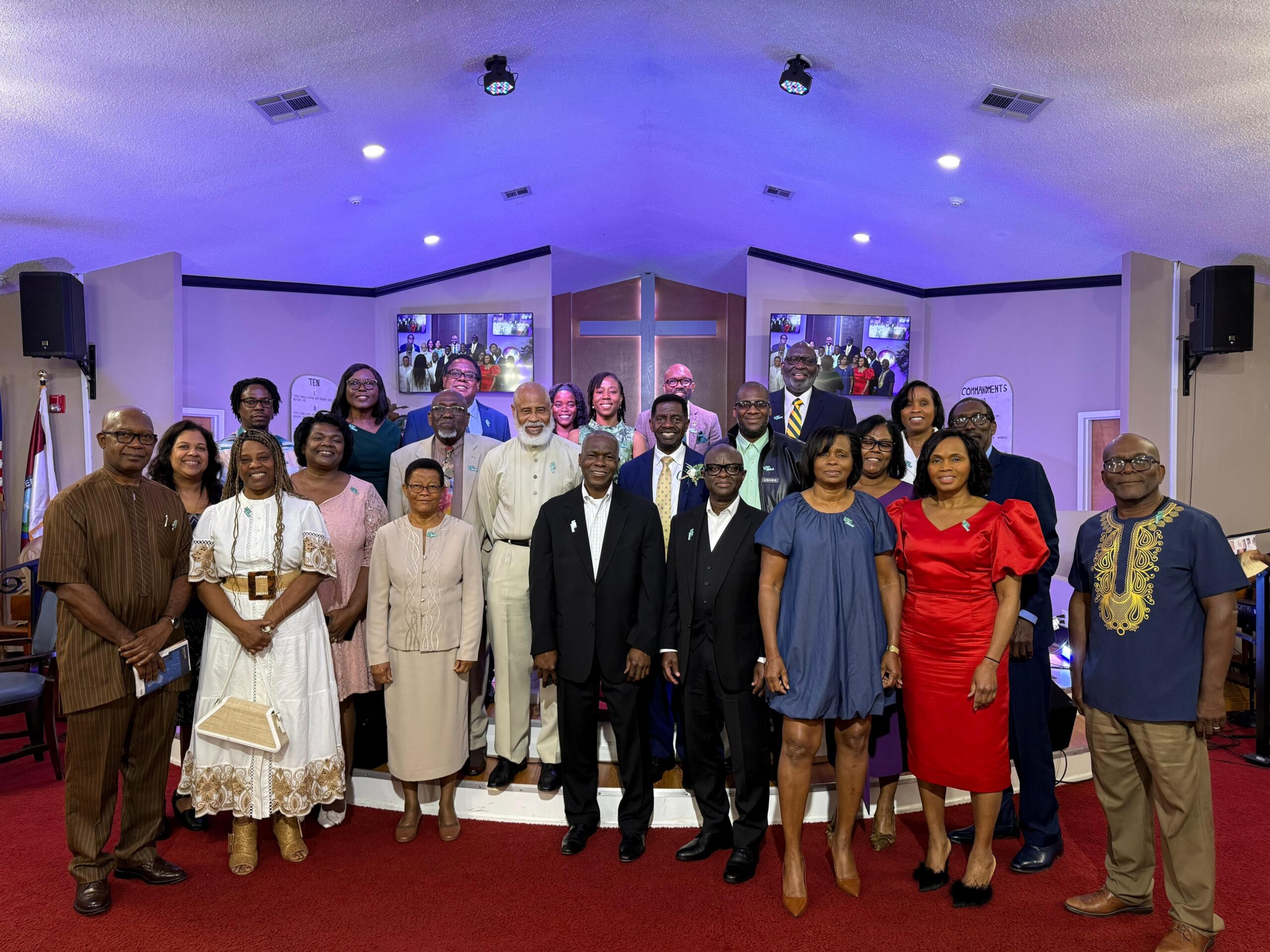 A large, smiling group of University of the Southern Caribbean alumni, family, and friends pose together for a group photograph inside Hebron Seventh-day Adventist Church in Texas.