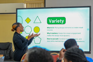 A female instructor stands in front of a large interactive display screen, gesturing with her hands to explain a presentation slide. The slide is titled "Variety" and features green and yellow geometric shapes alongside text defining the design concept. The backs of attendees' heads are visible in the foreground.