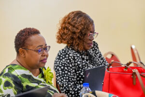 Two women seated at a seminar table, focusing attentively on their laptop screens. The woman on the left wears a green and white patterned top and glasses, while the woman slightly behind her wears a black and white polka dot top and glasses. A bright red handbag rests on the table.