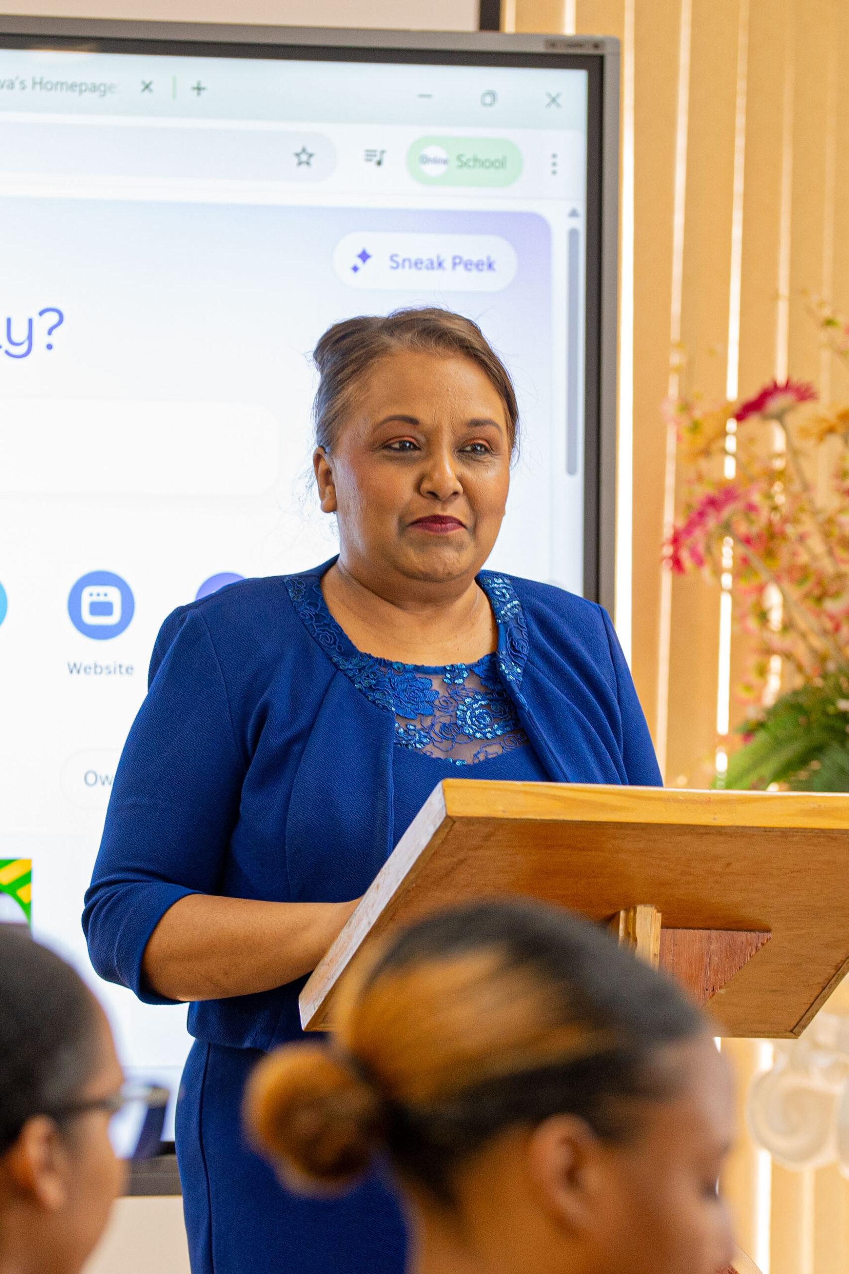 A woman, Mrs. Marsha Ramsamoojh-Harris, wearing a royal blue dress with lace detailing, stands behind a wooden podium addressing an audience. Behind her is a large digital screen displaying a web browser opened to Canva's homepage. The blurred heads of audience members are visible in the foreground.