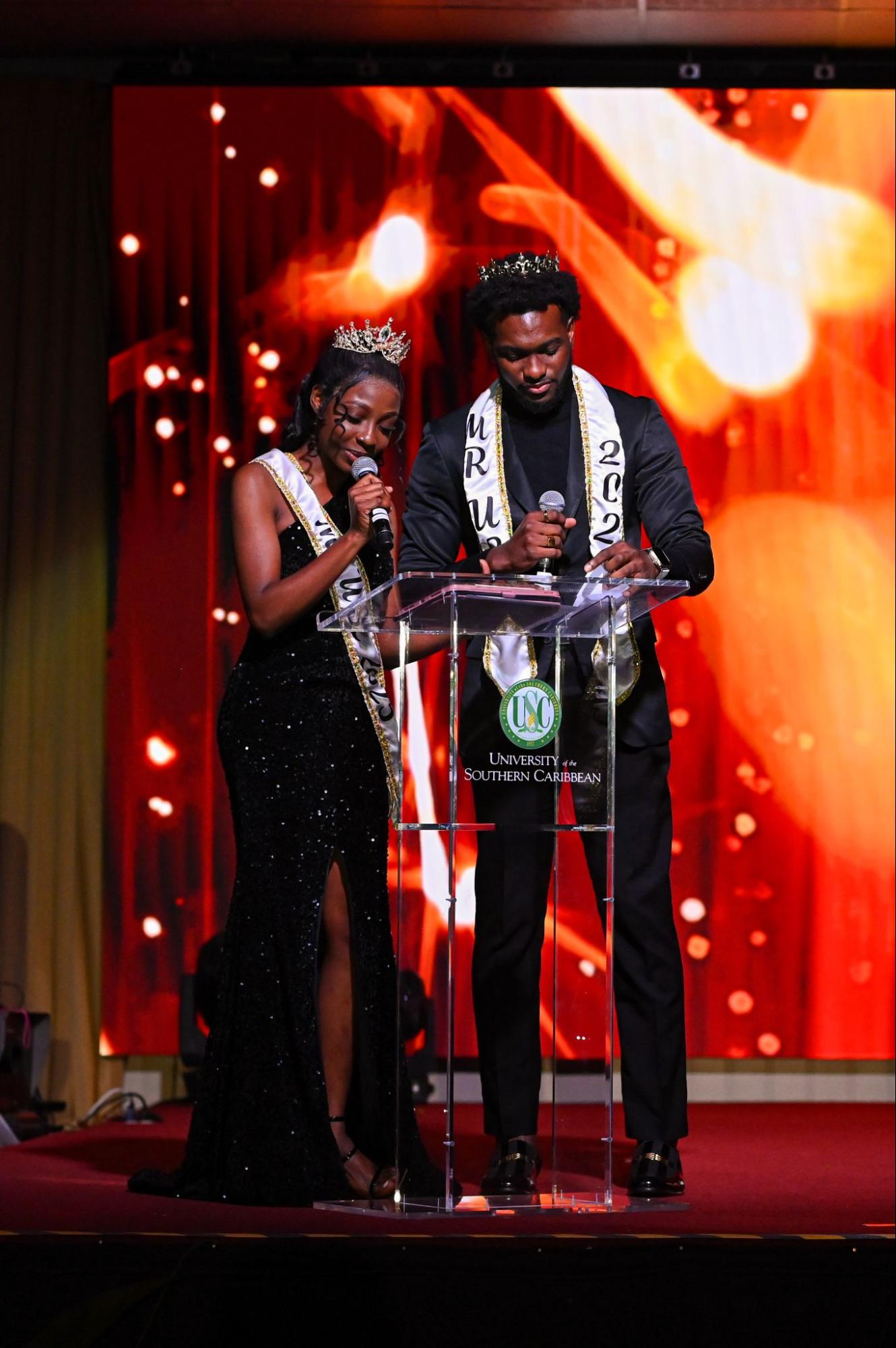 Outgoing titleholders Michael Garcia and Alicia Lewis standing at a transparent podium, speaking into microphones.