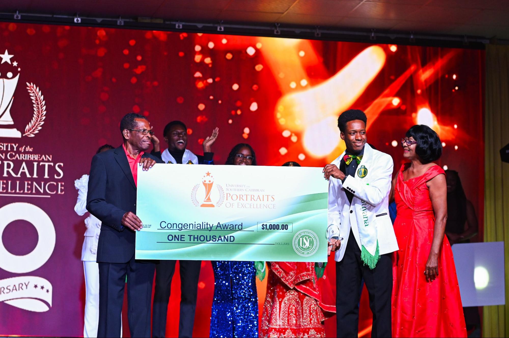 Adriell Wilson, wearing a white blazer, patterned bow tie, and green sash, smiles while receiving a large presentation cheque for one thousand dollars for the Congeniality Award.