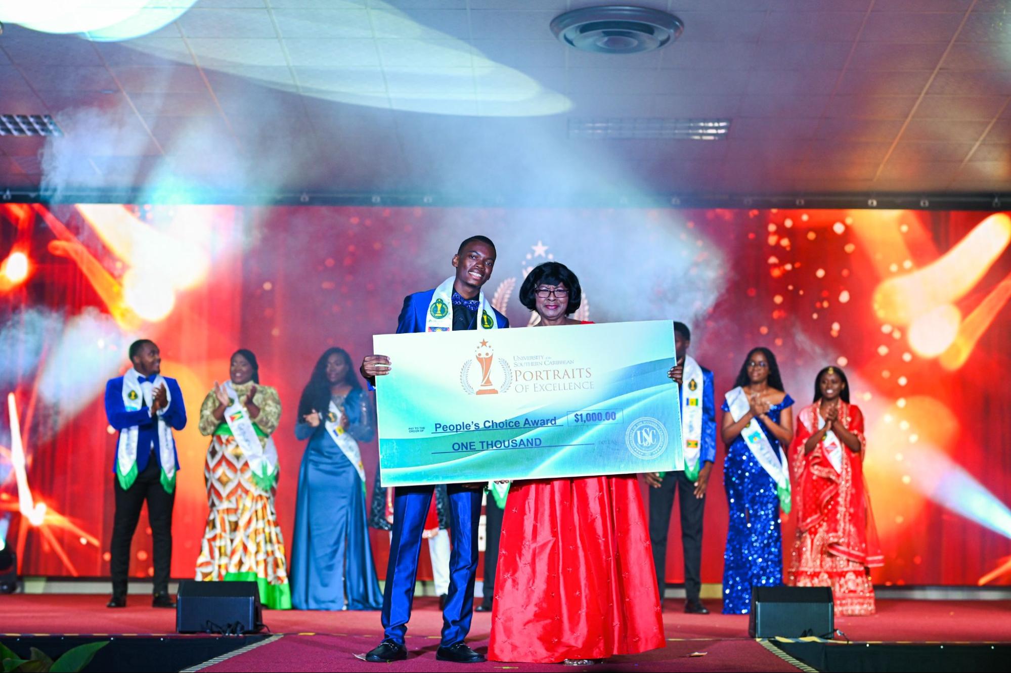 Lemario Clarke-Damier, dressed in a blue suit, holds a large presentation cheque for one thousand dollars for the People's Choice Award alongside a presenter in a red gown.