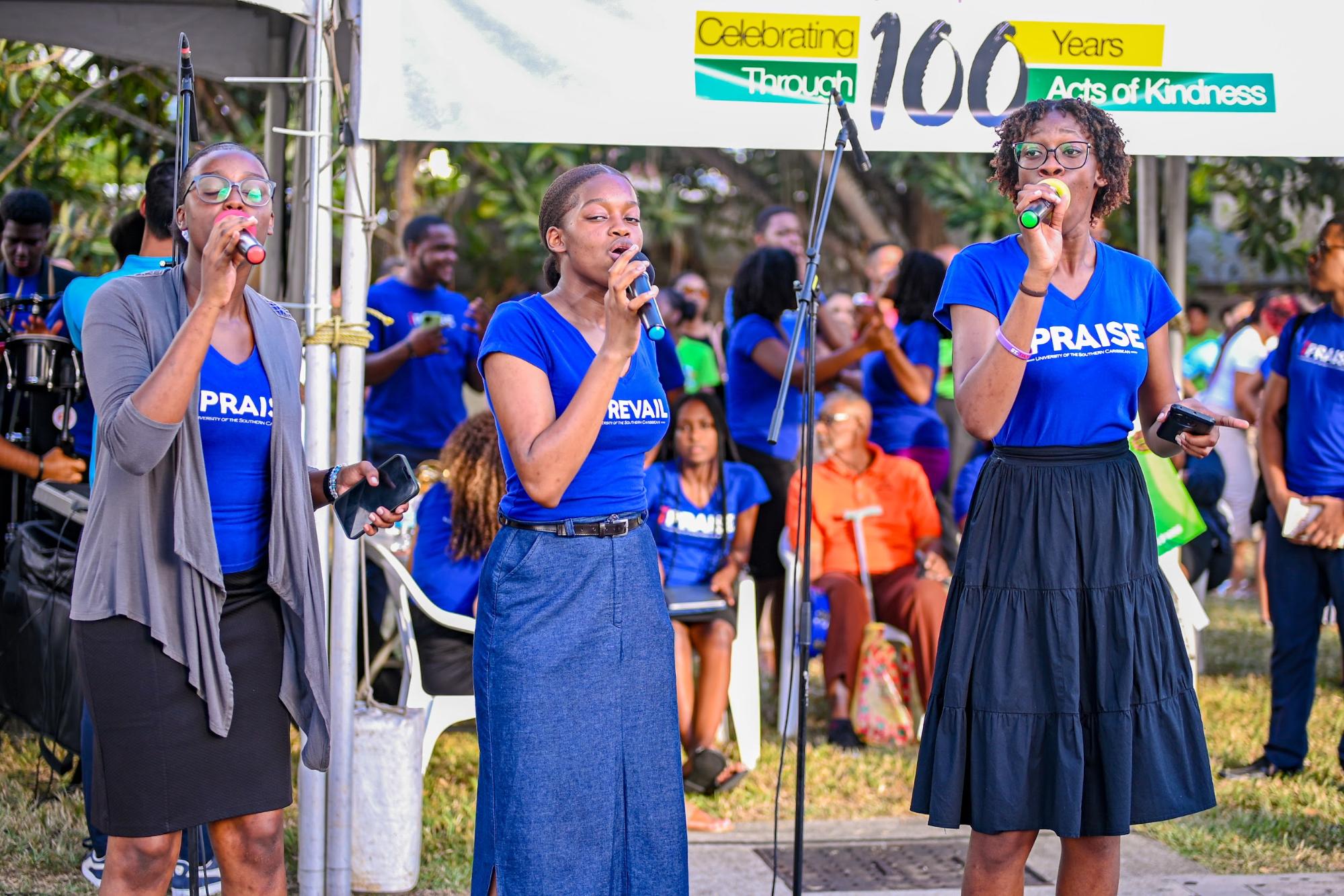 A group of young female students from the University of the Southern Caribbean singing passionately into microphones during the AOK-100 open-air launch event at Centre City Mall, Chaguanas