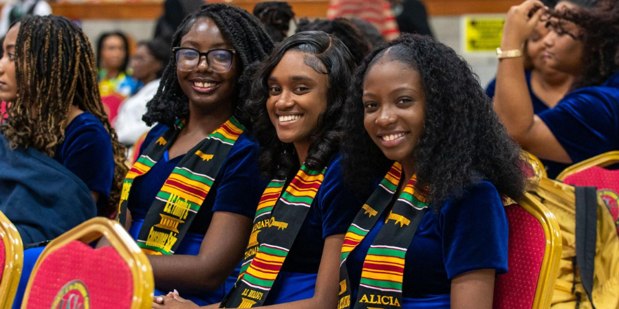 Three smiling female graduands sitting in red auditorium chairs, wearing matching blue velvet dresses and colourful graduation stoles, with Alicia Lewis on the far right.