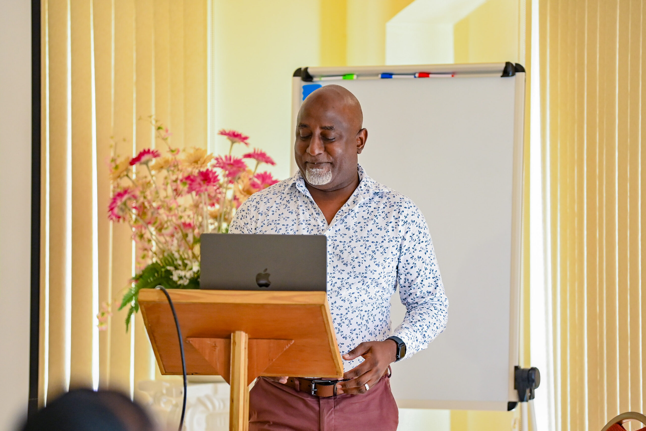 A man with a grey goatee, wearing a white patterned shirt, stands at a wooden podium. He is looking down at a dark grey laptop while delivering a presentation, with a floral arrangement visible in the background.