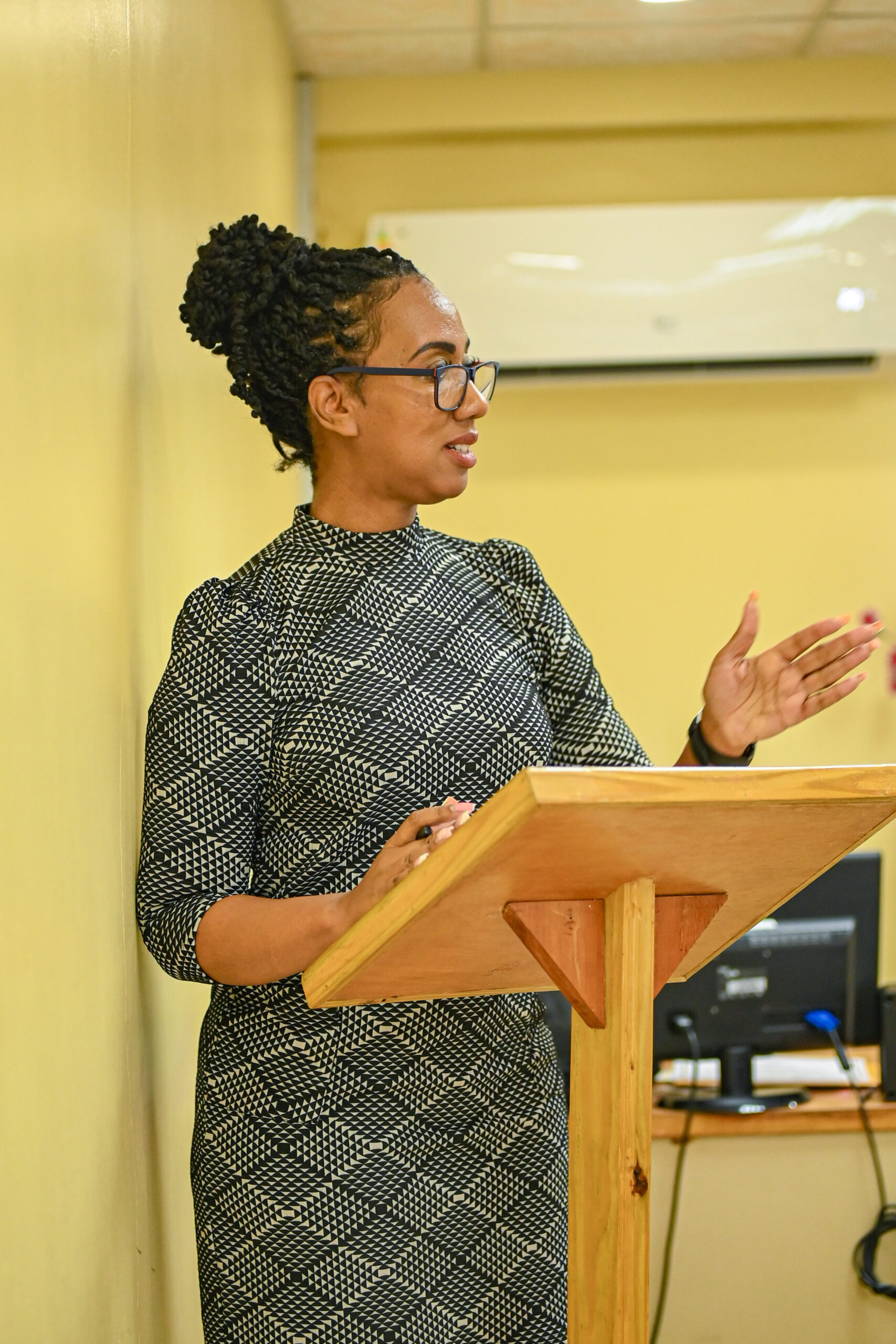 A woman with styled braided hair, wearing glasses and a black-and-white geometric patterned dress, speaks from behind a wooden podium. She is gesturing outward with her left hand as she presents.
