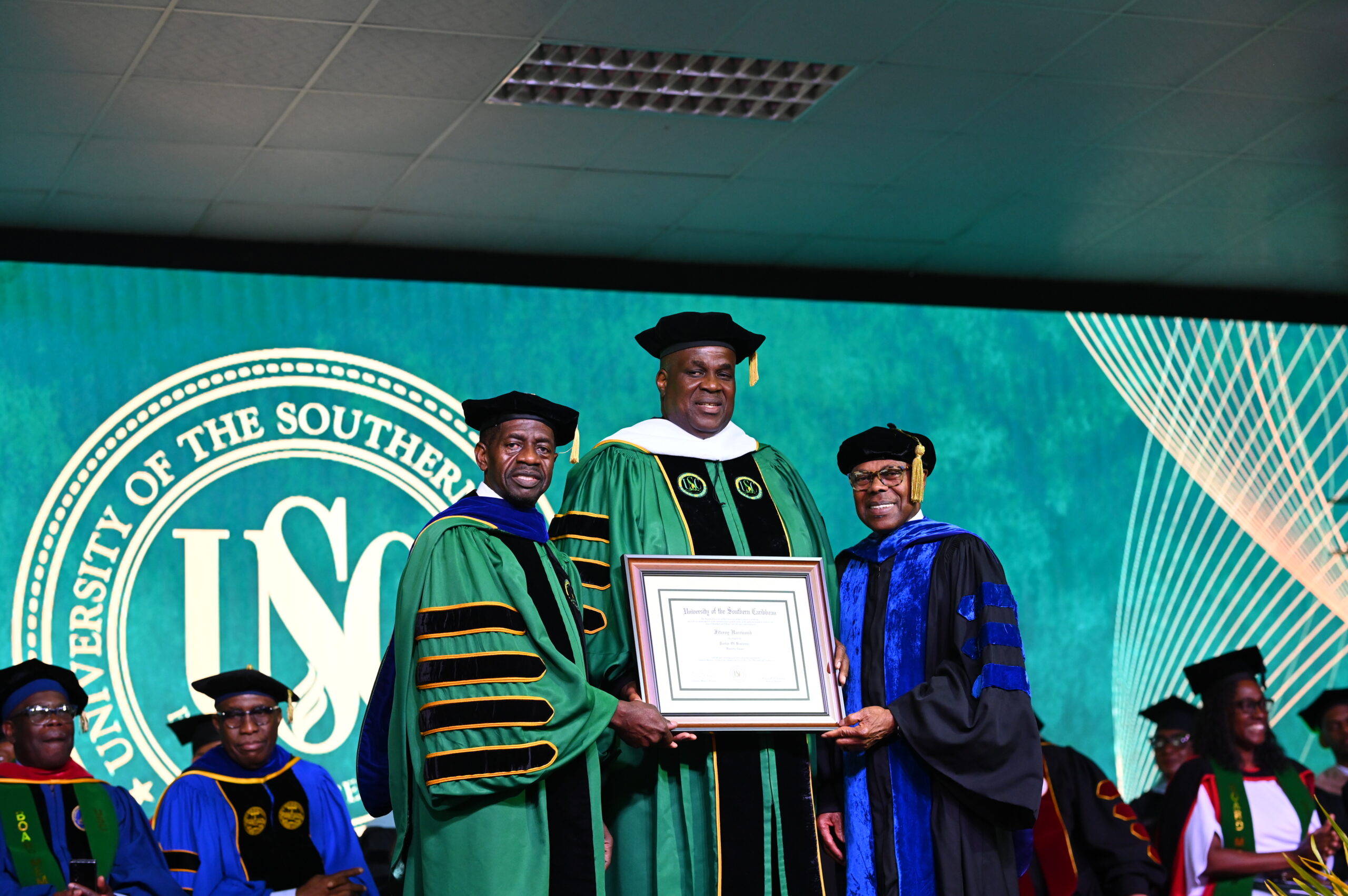 Mr Fitzroy Harewood stands in the centre holding his honourary degree certificate, flanked by Dr Colwick Wilson on the left and Dr Len Archer on the right in academic regalia.