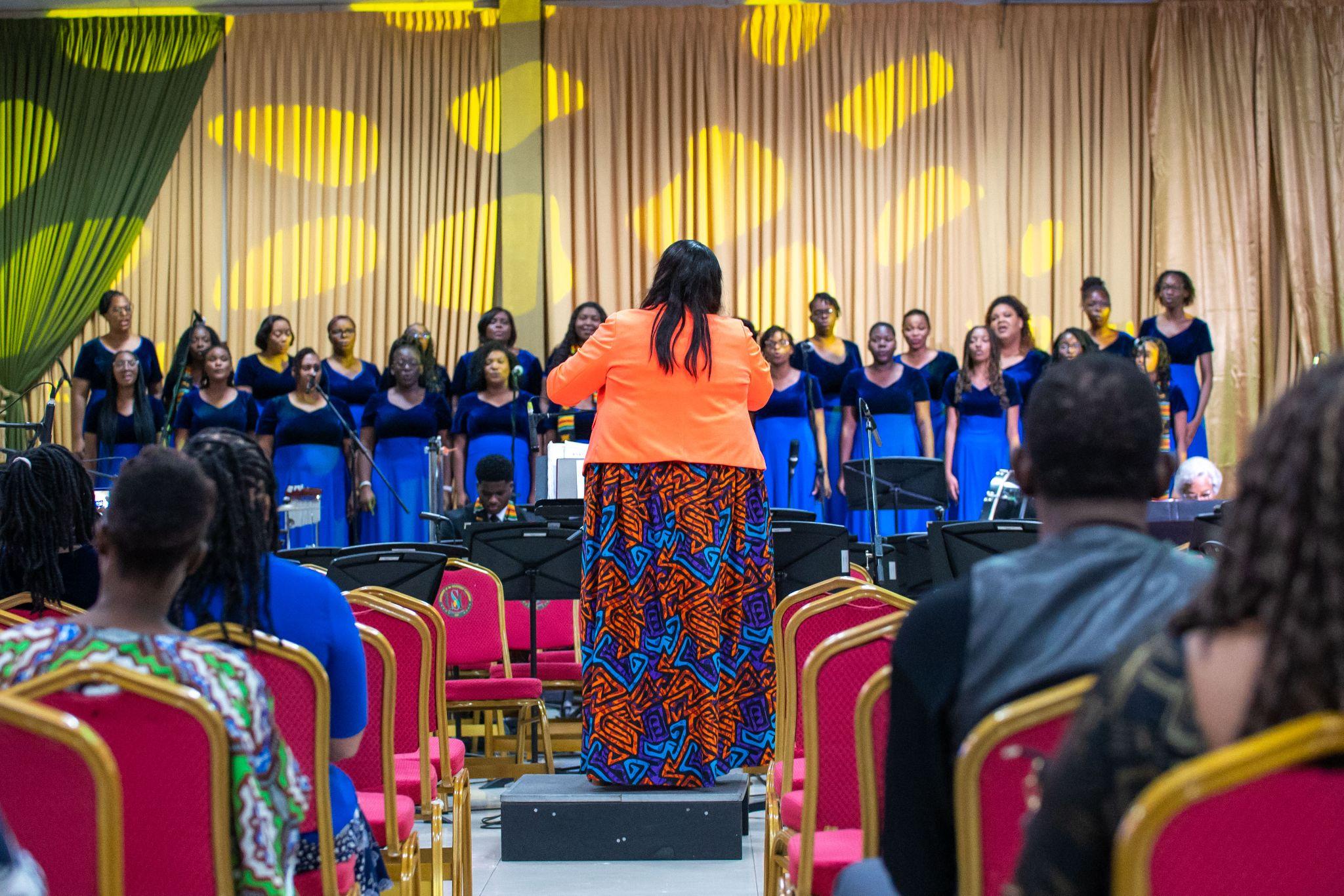 A choir conductor in a vibrant orange blazer and patterned skirt stands on a podium, facing away from the camera as she directs the USC Women’s Chorus on stage.