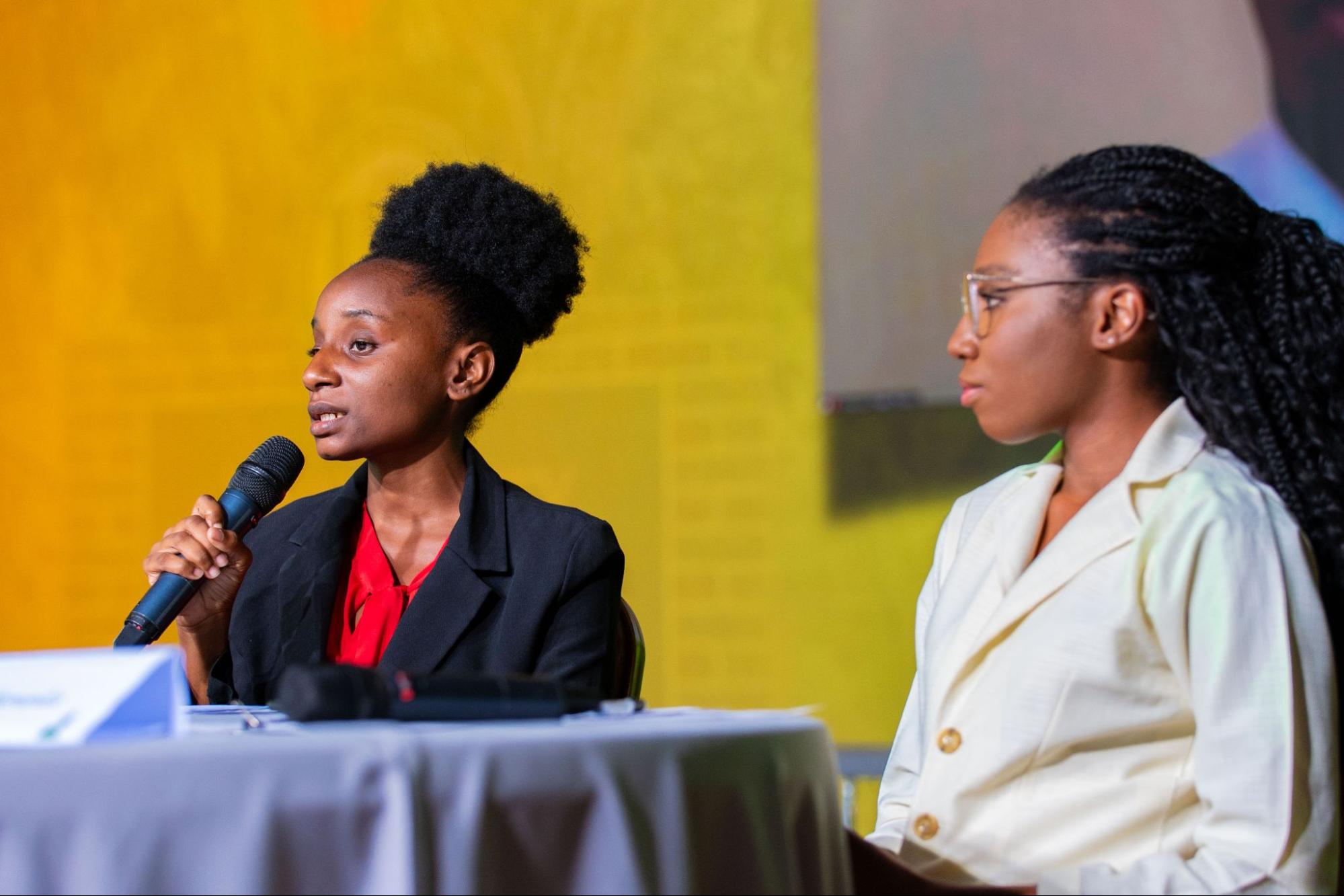 Elisa Farrell, VP candidate for Team Impact, speaks into a microphone while Presidential candidate Kalisia Hernandez sits to her right, looking toward the audience.