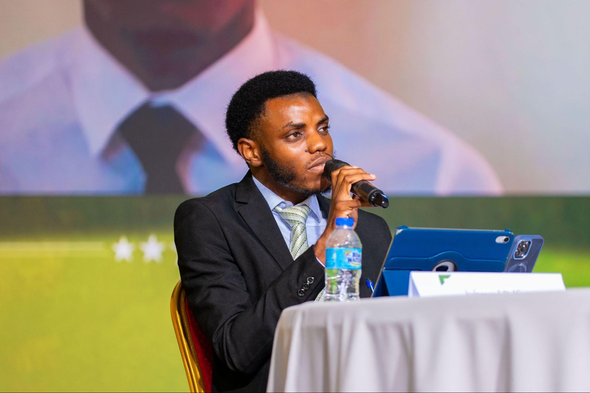 Presidential candidate Juhmual Belfon speaks into a microphone at a table with a tablet and water bottle. His Vice-Presidential candidate, Lemario De Clarke, is visible on a large screen in the background.