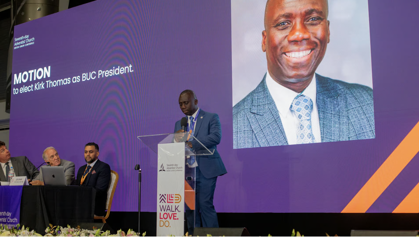 Dr Kirk Thomas speaks at a clear acrylic podium on a stage, with a massive purple digital screen behind him displaying his photo and the text "MOTION to elect Kirk Thomas as BUC President."