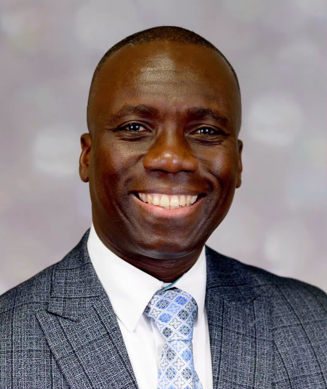 A professional portrait of Dr Kirk Thomas smiling warmly, dressed in a grey textured suit jacket, white shirt, and a light blue patterned tie.