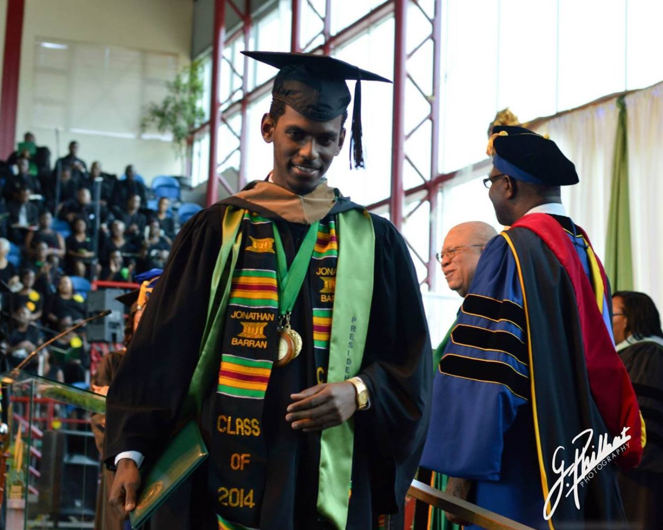 Jonathan Barran smiles broadly during his university graduation, wearing academic regalia that includes a green stole, a 'Class of 2014' sash, and an Associated Student Body President sash.