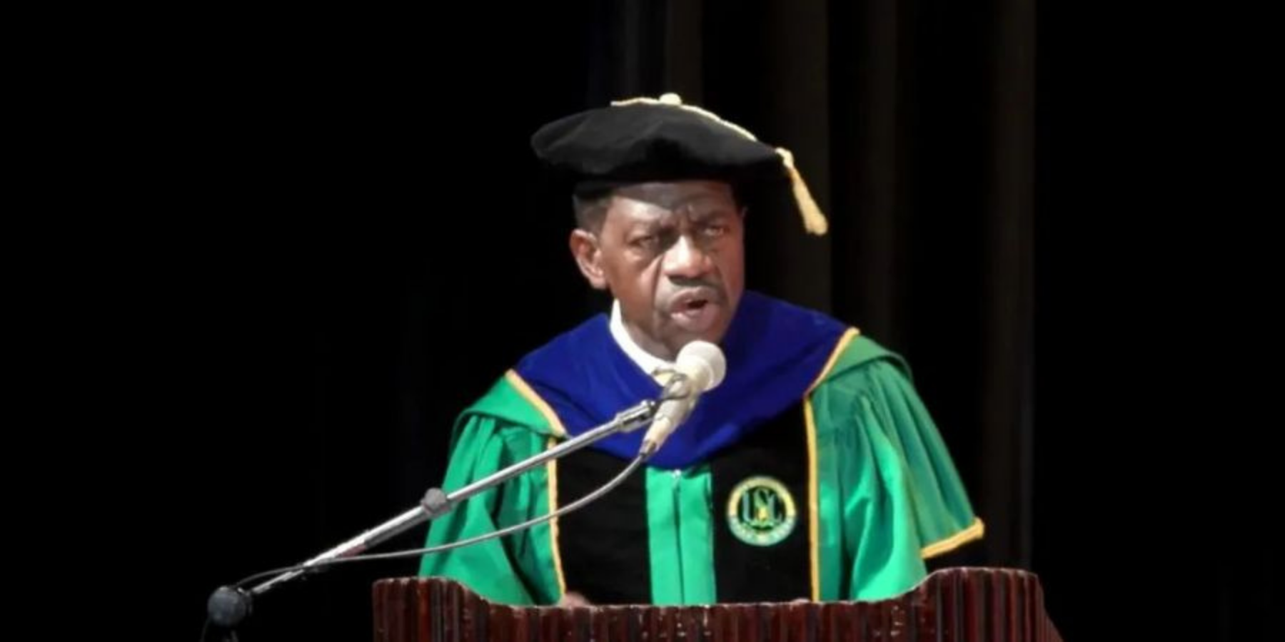 A medium close-up of Dr. Colwick M. Wilson speaking behind a wooden podium into a microphone, dressed in green and black doctoral regalia featuring a blue hood and a black velvet tam with a gold tassel against a dark background.