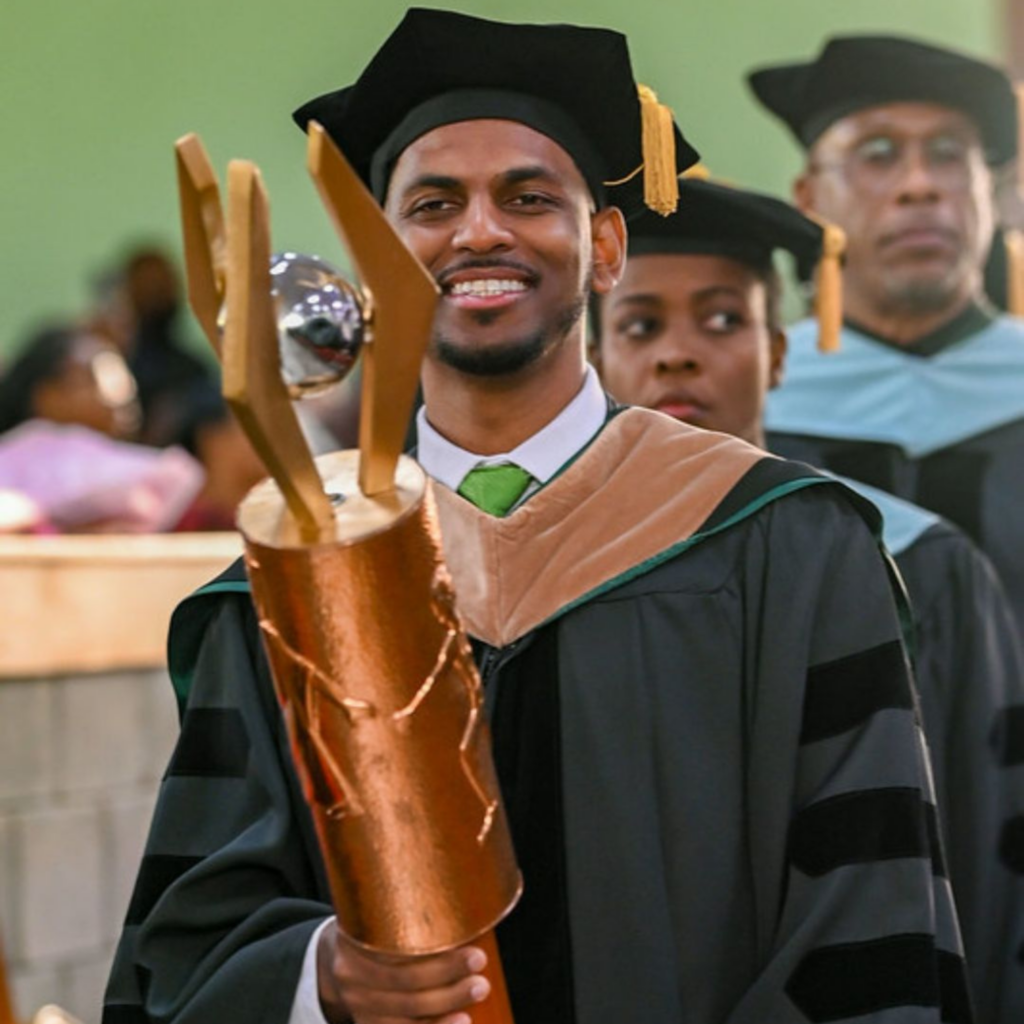 Dr Jonathan Barran smiles confidently in academic regalia, holding the ceremonial copper university mace as he leads the commencement procession.