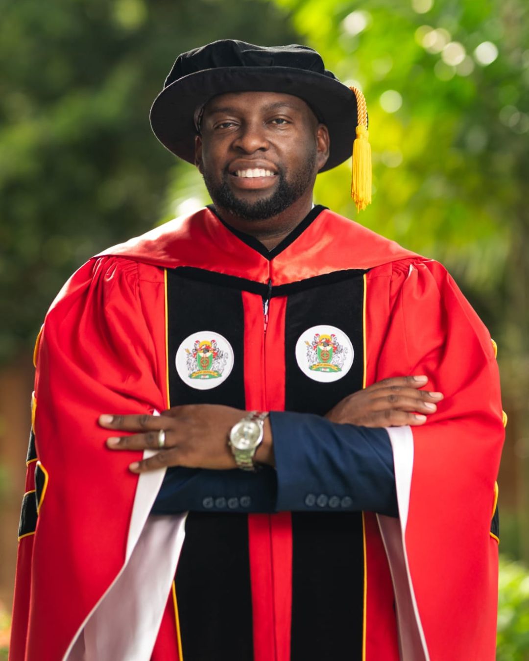 Dr Drason Browne smiles warmly with his arms crossed, wearing a bright red and black doctoral gown adorned with academic crests and a black velvet tam with a gold tassel.