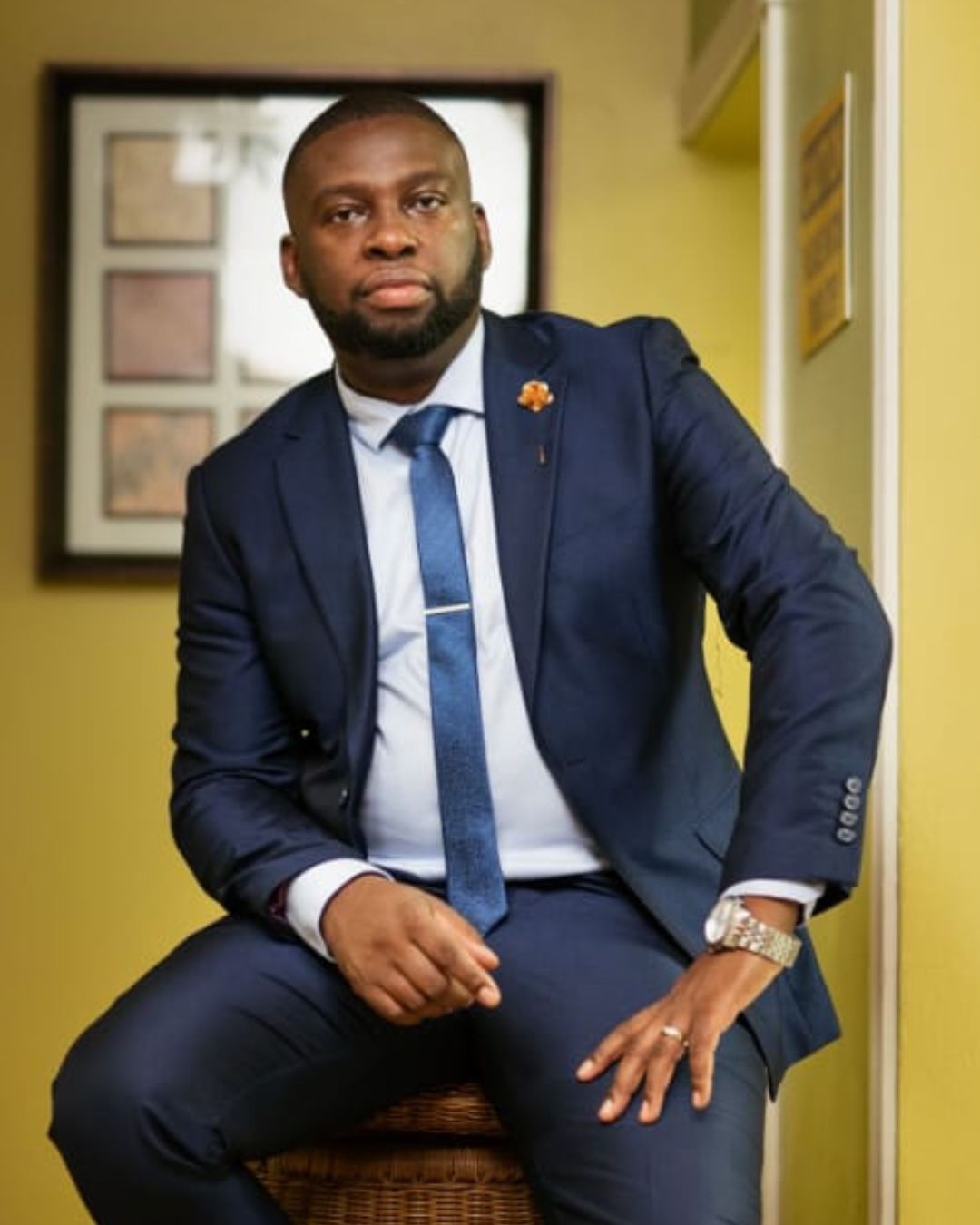 Dr Drason Browne sits confidently in a tailored navy blue suit and tie, looking directly at the camera while resting on a wicker stool in a brightly lit corridor.