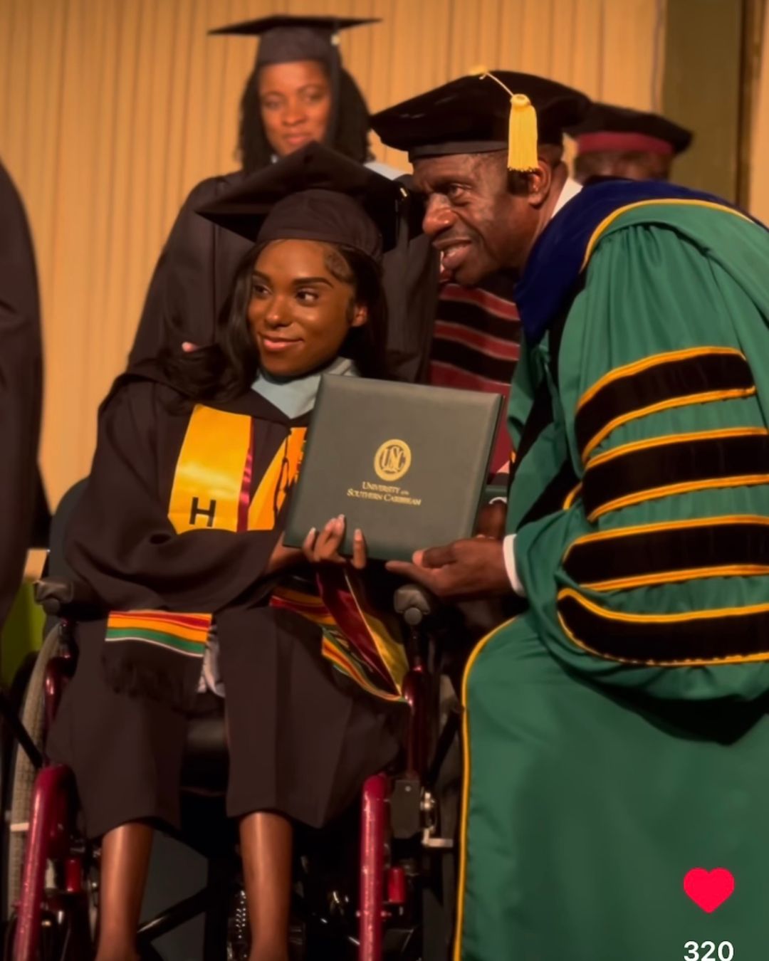 Gabrielle McKenzie, a young woman in a wheelchair wearing a black graduation gown and cap, smiles warmly as she receives her green diploma from a university official in green and black academic regalia on a commencement stage.
