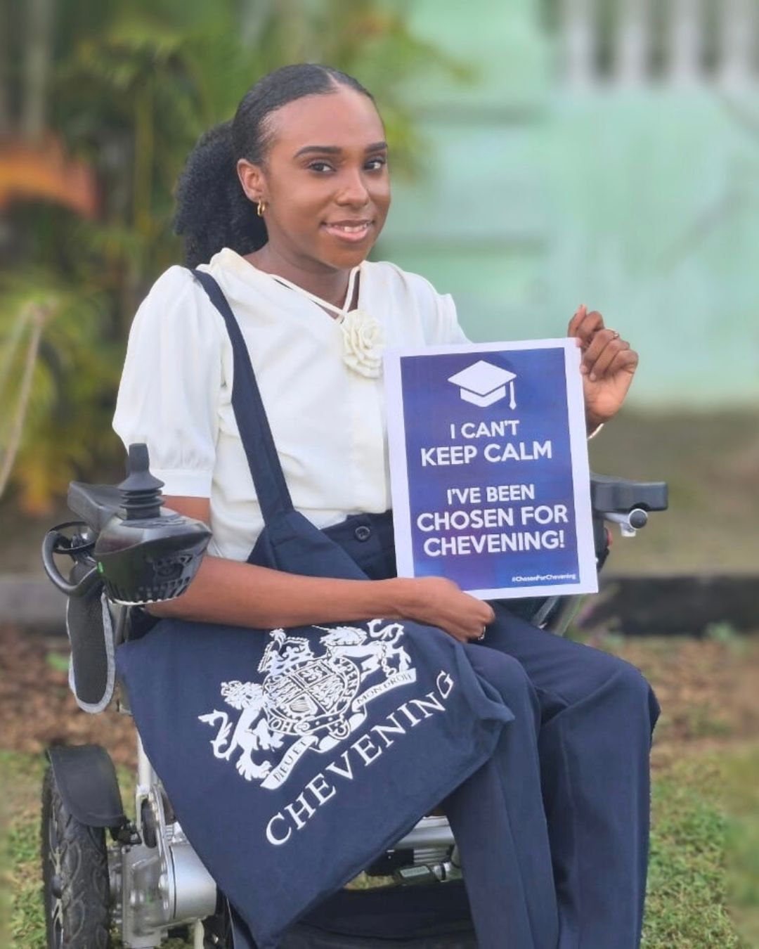 Gabrielle McKenzie sits outdoors in her wheelchair, smiling brightly. She is holding a printed sign that reads, "I CAN'T KEEP CALM I'VE BEEN CHOSEN FOR CHEVENING!" and has a navy blue Chevening-branded tote bag resting on her lap.