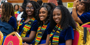 Three smiling female graduands sitting in red auditorium chairs, wearing matching blue velvet dresses and colourful graduation stoles, with Alicia Lewis on the far right.