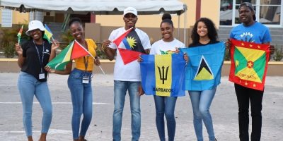 A diverse group of six smiling students at the University of the Southern Caribbean (USC) celebrating International Student Week. They are proudly displaying Caribbean flags, including the flags of Guyana, Antigua and Barbuda, Barbados, Saint Lucia, and Grenada, outdoors on the university campus.